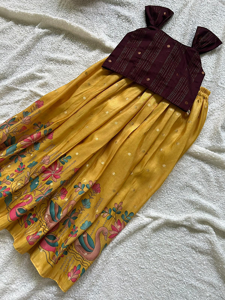 Flat lay of maroon sleeveless raw silk crop top with bow straps and pleated golden yellow skirt