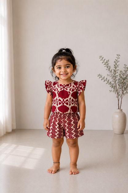 A young girl wearing the Adhira maroon kids cotton co-ord set featuring a flutter-sleeve top and zig-zag printed shorts.