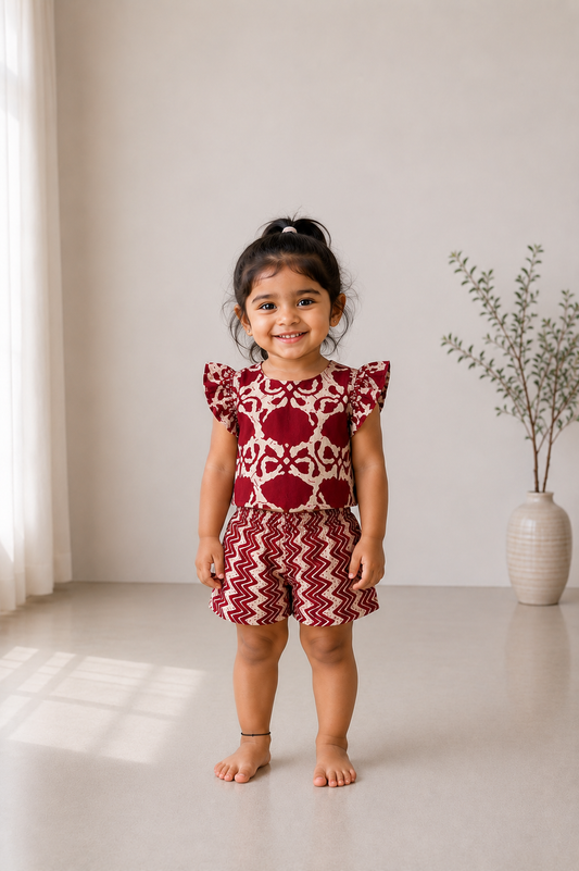 A young girl wearing the Adhira maroon kids cotton co-ord set featuring a flutter-sleeve top and zig-zag printed shorts.