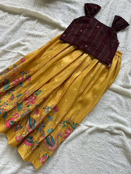 Flat lay of maroon sleeveless raw silk crop top with bow straps and pleated golden yellow skirt